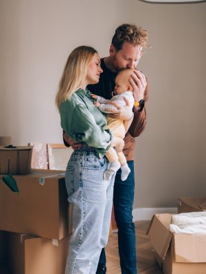 Man and woman holding a baby and standing in front of moving boxes